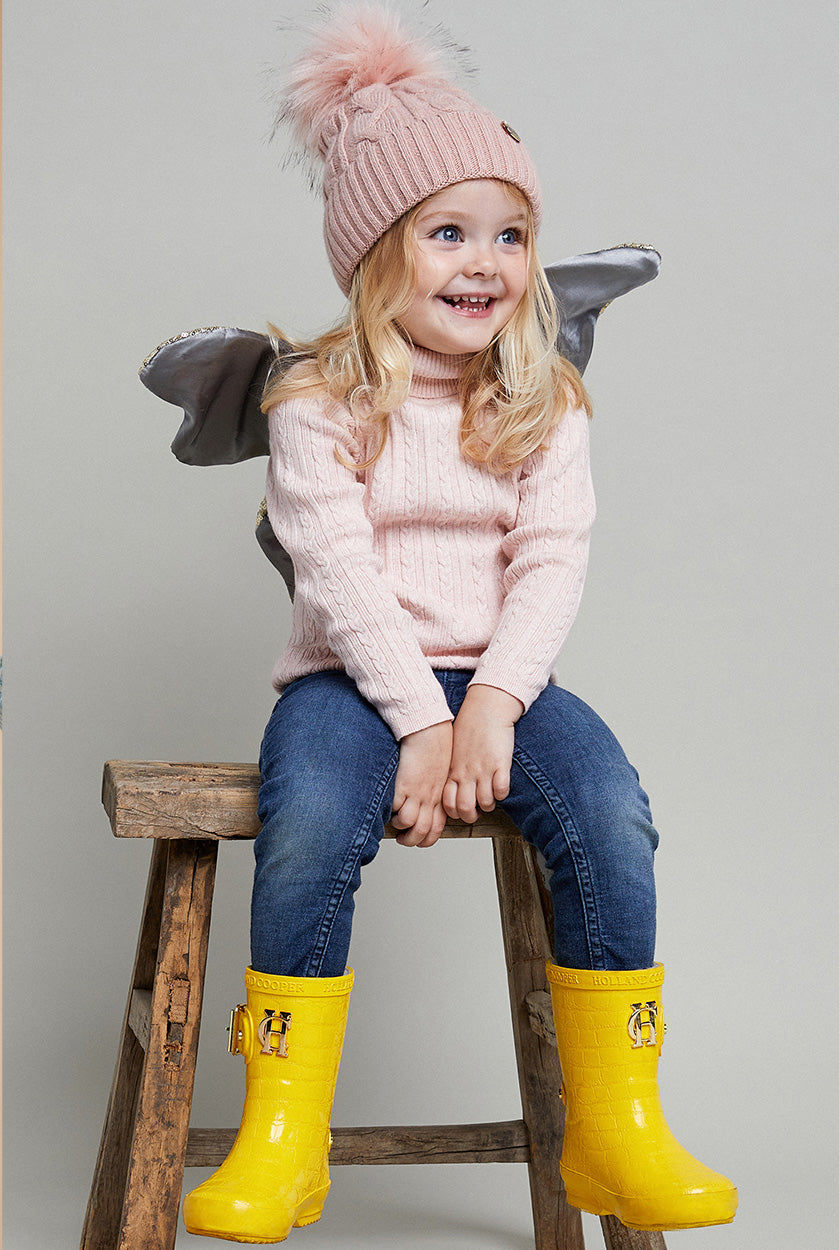Little blonde haired girl sat on wooden stool wearing pink jumper and jeans with yellow wellies and pink bobble hat